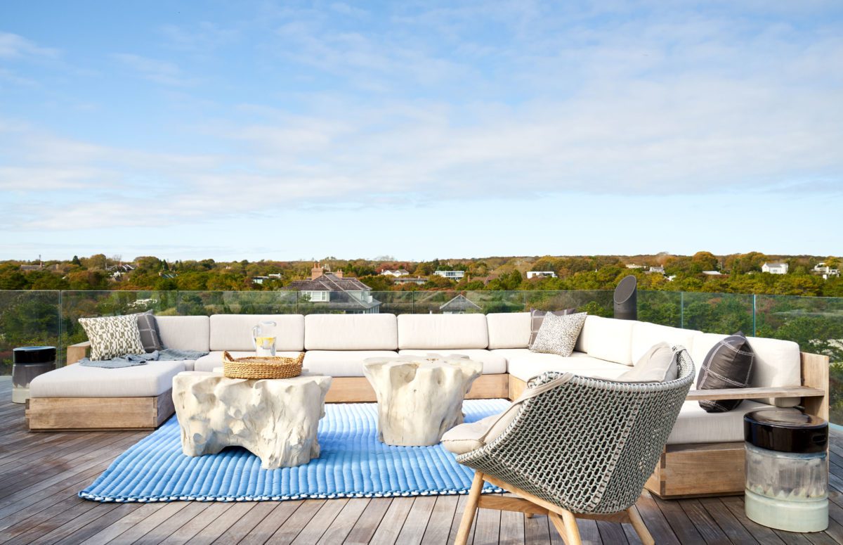 Outdoor view of rooftop lounge with crystal railing, a comfortable white sofa, and armchairs, sculptural tables.